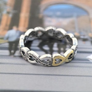 May include: A silver-toned ring with an infinity symbol design. The ring features a repeating pattern of interlocking loops, with a section of gold-toned metal. The ring is set against a blurred background.
