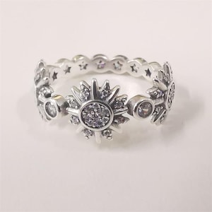 May include: A silver ring with a sunburst design, a central cluster of small, clear stones, and a band with star-shaped cutouts. The ring also features additional clear stones. The ring is approximately a size O.
