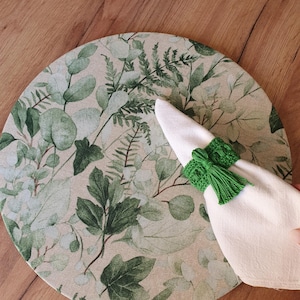 May include: A round placemat with a green leafy pattern. A white napkin is placed on top of the placemat and held in place with a green tassel ring.