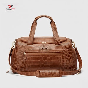 May include: Brown leather duffel bag with a crocodile skin texture. The bag has a top handle, a shoulder strap, and multiple zippered compartments. The bag has gold-colored hardware and the brand name "ROMFIT SPORTS" is visible in the upper left corner.