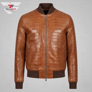Puede incluir: Chaqueta bomber de cuero marrón con textura de piel de cocodrilo. La chaqueta tiene cierre de cremallera, dos bolsillos laterales y puños y dobladillo de canalé marrón. El cuello es de color marrón oscuro. La chaqueta es de color marrón medio.