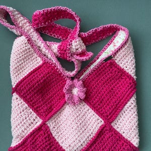 Könnte beinhalten: Handgefertigte Häkeltasche in Rosa- und Hellrosa-Tönen mit einem Rautenmuster. Die Tasche hat einen passenden Träger und eine dekorative Blume. Das Innenfutter hat ein Blumenmuster.