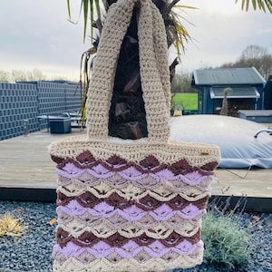 Könnte beinhalten: Handgefertigte Häkeltasche in Beige-, Braun- und Lavendeltönen. Die Tasche hat ein Muschelmuster am Körper und einen einfarbigen beigen Griff. Die Tasche wird im Freien an einer Palme befestigt gezeigt.