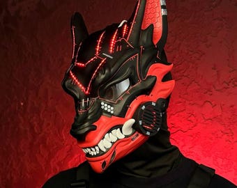 May include: A black and red cybernetic wolf mask with glowing red accents. The mask features sharp teeth and a detailed design. The person is wearing a black tactical vest with a white and black patch.