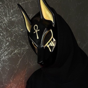 Puede incluir: Una máscara negra de Anubis con detalles dorados en las orejas y los ojos, con un símbolo ankh. La máscara se usa con una sudadera con capucha negra, creando una apariencia misteriosa y llamativa.