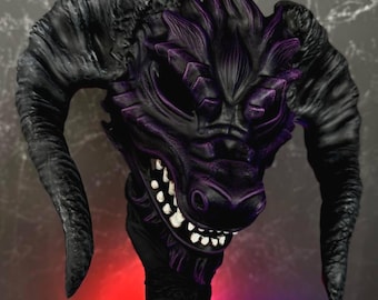 May include: A black and purple dragon mask with large curved horns and sharp teeth. The mask is held up against a dark background with red and purple lighting. The mask has a detailed, textured surface.