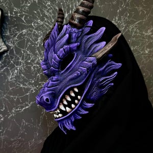Può includere: Una maschera di drago viola dettagliata con denti affilati, corna e squame strutturate. La maschera è indossata sopra un indumento nero, creando un contrasto sorprendente. Il design della maschera è intricato, con un focus sul volto del drago.