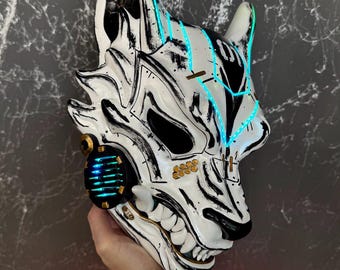 May include: A white and black wolf mask with neon blue accents. The mask features a detailed design with gold and black accents, and a glowing blue light on the side. The mask is held up by a hand.