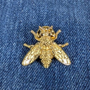 Puede incluir: Broche dorada con forma de abeja. El broche tiene alas detalladas, segmentos corporales y antenas. La abeja está sobre un fondo de mezclilla azul.