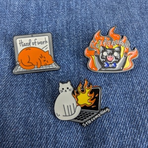 May include: Three enamel pins with cat designs on a denim background. One pin shows an orange cat sleeping on a laptop with the text "Hard of work." Another shows a cat in flames with the text "Let it burn!" The third shows a white cat with a laptop on fire and the text "Still compiling."