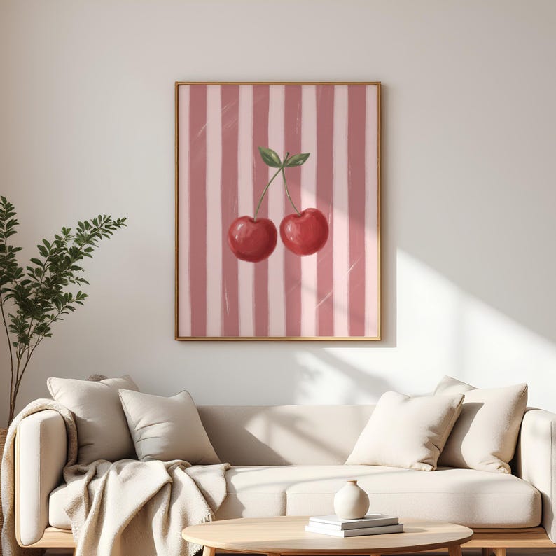 May include: A framed art print featuring two red cherries with green leaves against a pink and white striped background. The artwork is displayed above a beige sofa with pillows and a blanket.