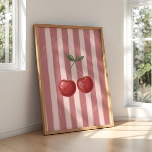 May include: A framed art print featuring two red cherries with green leaves against a pink and white striped background. The artwork is displayed in a light brown wooden frame, leaning against a white wall.