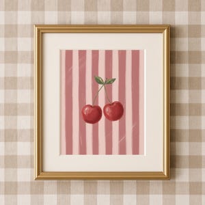 May include: A framed art print featuring a pair of ripe red cherries with green leaves against a pink and white striped background. The artwork is set in a gold frame with a white mat, suitable for kitchen or dining room decor.
