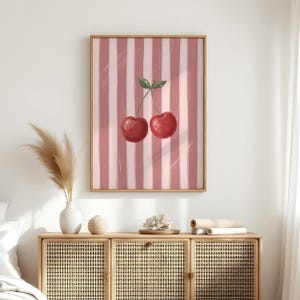 May include: A framed art print featuring two ripe red cherries with green leaves against a pink and white striped background. The artwork is displayed in a gold-colored frame, adding a touch of elegance to the fruit-themed illustration.