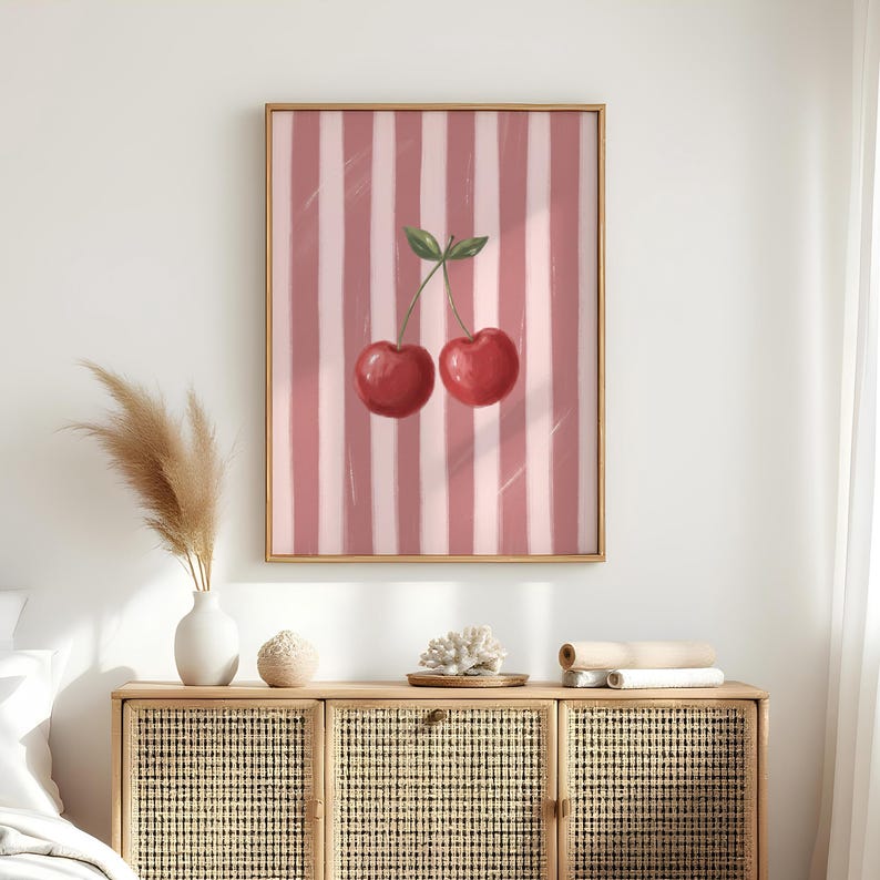 May include: A framed art print featuring two ripe red cherries with green leaves against a pink and white striped background. The artwork is displayed in a gold-colored frame, adding a touch of elegance to the fruit-themed illustration.