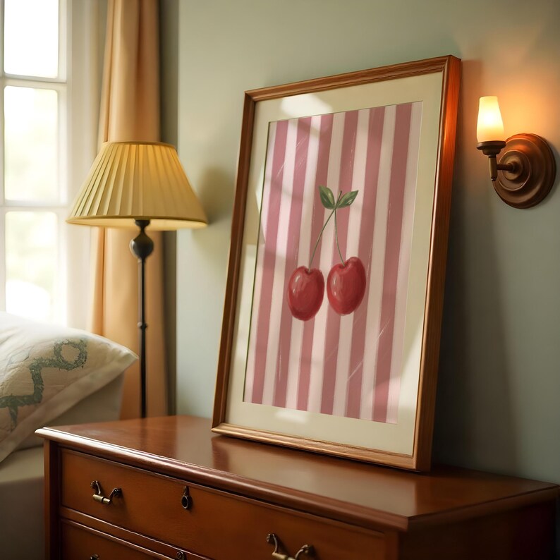 May include: A framed art print featuring two red cherries with green leaves against a pink and white striped background. The artwork is displayed on a wooden dresser. A yellow lamp and a wall sconce are also visible.
