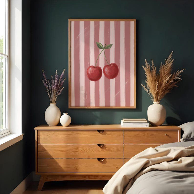 May include: A framed art print featuring two red cherries with green leaves against a pink and white striped background. The artwork is displayed above a wooden dresser with decorative vases and dried plants, creating a cozy interior design.