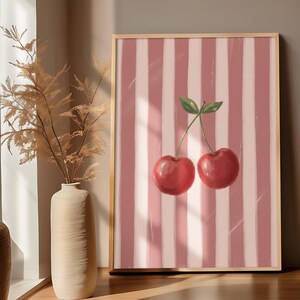 May include: A framed art print featuring two red cherries with green leaves against a pink and white striped background. The artwork is in a wooden frame and is displayed indoors.
