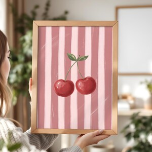 May include: A framed art print featuring a pair of red cherries with green leaves against a pink and white striped background. The artwork is displayed in a light brown wooden frame, suitable for kitchen or dining room decor.