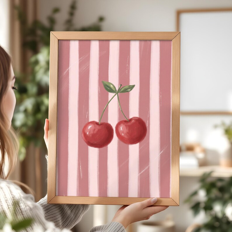 May include: A framed art print featuring a pair of red cherries with green leaves against a pink and white striped background. The artwork is displayed in a light brown wooden frame, suitable for kitchen or dining room decor.