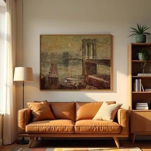 Op de afbeelding: Een ingelijste vintage schilderij van een brug over water, waarschijnlijk de Brooklyn Bridge, hangt boven een bruine leren bank. Het schilderij heeft een gedempt kleurenpalet en zit in een houten lijst. Een vloerlamp en een houten boekenkast staan ook in de kamer.