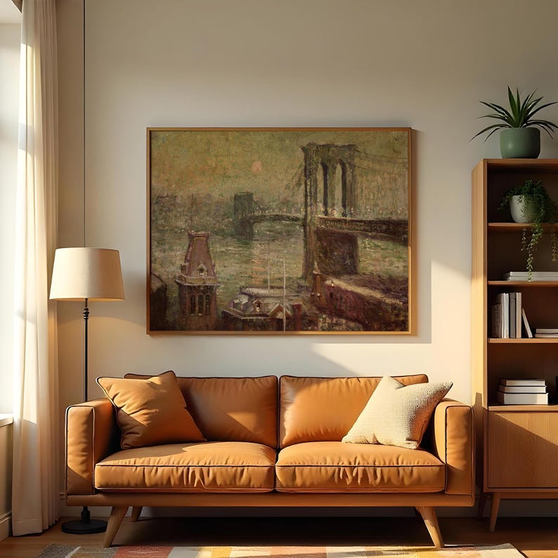 Op de afbeelding: Een ingelijste vintage schilderij van een brug over water, waarschijnlijk de Brooklyn Bridge, hangt boven een bruine leren bank. Het schilderij heeft een gedempt kleurenpalet en zit in een houten lijst. Een vloerlamp en een houten boekenkast staan ook in de kamer.
