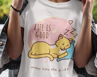 Puede incluir: Camiseta blanca con un gato amarillo durmiendo sobre una almohada azul, con el texto "LIFE IS GOOD WHEN YOU ARE A CAT". El gato está frente a un círculo rosa con un corazón y Zzz.