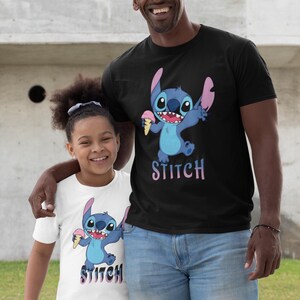 Op de afbeelding: Zwart T-shirt met een blauw stripfiguur dat ijs vasthoudt, met het woord "STITCH" in roze en paars. Een wit T-shirt met hetzelfde ontwerp is ook zichtbaar.