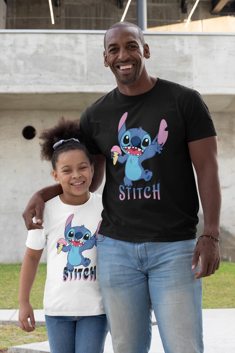 Op de afbeelding: Zwart T-shirt met een blauw stripfiguur dat ijs vasthoudt, met het woord "STITCH" in roze en paars. Een wit T-shirt met hetzelfde ontwerp is ook zichtbaar.