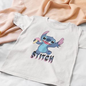 Op de afbeelding: Wit T-shirt met een cartoon illustratie van Stitch, een blauwe alien, die ijs vasthoudt. Het woord "Stitch" staat eronder in paars en zwart. Het shirt ligt op een witte ondergrond.