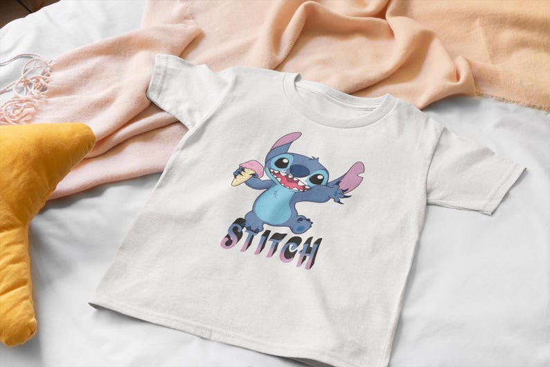 Op de afbeelding: Wit T-shirt met een cartoon illustratie van Stitch, een blauwe alien, die ijs vasthoudt. Het woord "Stitch" staat eronder in paars en zwart. Het shirt ligt op een witte ondergrond.