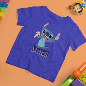 Op de afbeelding: Een blauw t-shirt met het personage Stitch met een ijsje. Het woord "Stitch" staat eronder. De afbeelding is omgeven door kinderspeelgoed, waaronder een xylofoon, een basketbal en een speelgoedauto.