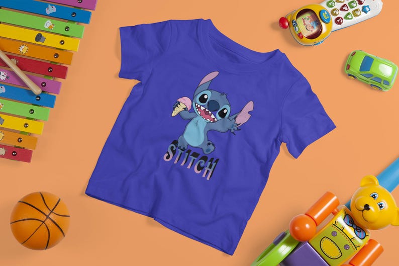 Op de afbeelding: Een blauw t-shirt met het personage Stitch met een ijsje. Het woord "Stitch" staat eronder. De afbeelding is omgeven door kinderspeelgoed, waaronder een xylofoon, een basketbal en een speelgoedauto.