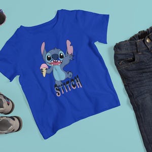 Op de afbeelding: Een blauw t-shirt met een cartoon Stitch-personage dat ijs vasthoudt, met het woord "Stitch" eronder. De afbeelding bevat ook een blauwe jeans, een zonnebril en kindersandalen.