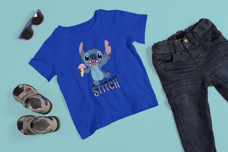 Op de afbeelding: Een blauw t-shirt met een cartoon Stitch-personage dat ijs vasthoudt, met het woord "Stitch" eronder. De afbeelding bevat ook een blauwe jeans, een zonnebril en kindersandalen.