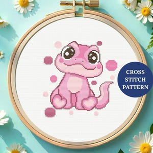 Könnte beinhalten: Ein Kreuzstichmuster mit einer rosa Eidechse mit großen Augen und rosa Punkten. Das Design befindet sich in einem hölzernen Stickrahmen. Die Worte "CROSS STITCH PATTERN" stehen auf einem blauen Kreis. Gänseblümchen und Blätter umgeben den Rahmen.