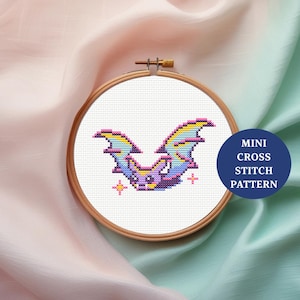May include: A cross-stitch bat design in a wooden embroidery hoop, featuring purple, blue, yellow, and pink hues. The text "MINI CROSS STITCH PATTERN" is on a blue circle. The design is on white fabric.