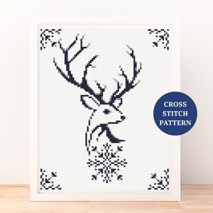 May include: A framed cross-stitch pattern featuring a black and white deer head with large antlers and a snowflake. The design is set against a white background with decorative corner accents. A blue circle with the text "CROSS STITCH PATTERN" is in the lower right corner.