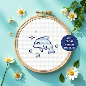 Może przedstawiać: Wzór haftu krzyżykowego przedstawiający delfina w drewnianym tamborku. Delfin jest jasnoniebieski z ciemnoniebieskimi akcentami, otoczony ciemnoniebieskimi bąbelkami i gwiazdami. Granatowe koło z napisem "MINI CROSS STITCH PATTERN" znajduje się po prawej stronie.