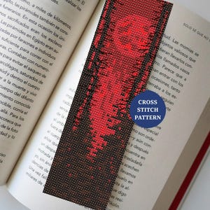 Pode incluir: Um marcador de livros feito à mão com um padrão de ponto cruz em vermelho e preto, pousado em um livro aberto. O marcador de livros apresenta um design gradiente. O texto "CROSS STITCH PATTERN" está em um círculo azul.