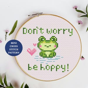 May include: A mini cross stitch pattern in a wooden hoop. The design features a green frog with the text "Don't worry, be hoppy!" and a pink heart. The background is light pink.