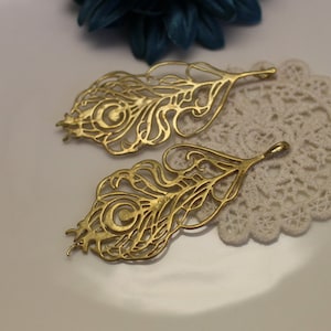 May include: Two gold-colored metal peacock feather charms with intricate cut-out designs. The feathers are delicate and have a slightly textured surface.