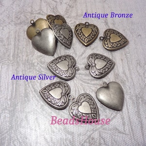 May include: Antique silver and bronze heart-shaped locket charms with floral and bow details. The lockets are open and have a smooth interior.