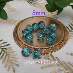 Pode incluir: Close-up de contas de vidro azul turquesa de 8 mm com design facetado. As contas estão dispostas em um prato dourado. O texto "8mm" e "BeadsHouse" é visível na imagem.