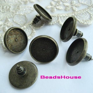 May include: Five antique bronze-colored metal earring settings with round, flat tops. The settings have a post on the back for attaching earrings.
