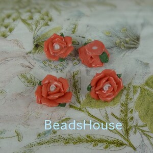 May include: Four coral-colored rose-shaped beads with white centers and green leaves are arranged on a floral patterned background. The text "BeadsHouse" is visible at the bottom of the image. These beads are suitable for jewelry making.