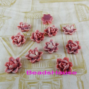 Pode incluir: Dez cabochons de flores de resina rosa com um centro branco. As flores estão em várias posições sobre um tecido com padrão branco e dourado. O texto "BeadsHouse" é visível no centro inferior da imagem.