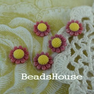 May include: Five pink and yellow flower-shaped buttons with a yellow center. The buttons are arranged on a white crocheted doily and a floral patterned fabric.