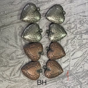 May include: Eight heart-shaped pendants in silver and copper tones. Each pendant has a textured surface with a decorative border and a small loop at the top. The pendants are arranged on a textured gray surface.