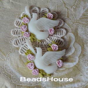 May include: Two white ceramic doves with pink roses and green leaves. The doves are facing each other and have their wings spread out. The roses are arranged around the doves in a circle.